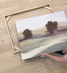 a woman holding up a painting in front of a wooden frame on the floor next to it