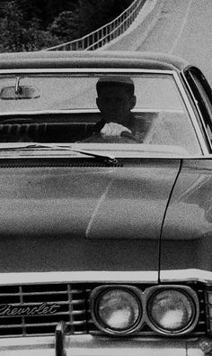 a black and white photo of a man driving a car down a road with another person in the back seat