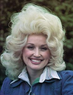 a woman with blonde hair smiling and wearing a blue jacket in front of some trees