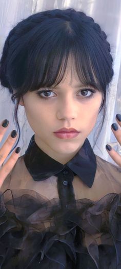 a woman with black hair and nails wearing a sheer shirt, holding her hands up to her face