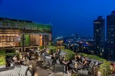 people sitting at tables on top of a roof with city lights in the back ground