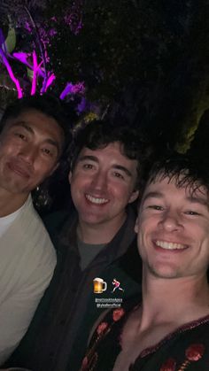 three men are smiling for the camera in front of some trees and bushes at night