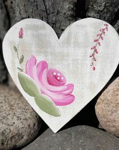 a white heart with pink flowers painted on it sitting next to some rocks and stones