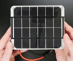 a hand holding a small solar panel with an orange cord attached to it and two hands on the side