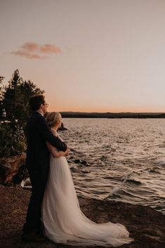 a bride and groom standing on the shore at sunset looking out over the water with their arms around each other