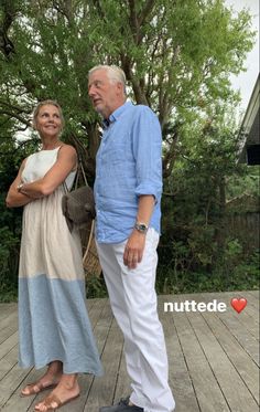 an older man and woman standing on a wooden deck in front of a tree with their arms crossed