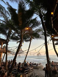 many people are sitting at tables on the beach near the water and some palm trees