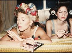 two women laying on a bed with hair dryers on their heads and one reading a book