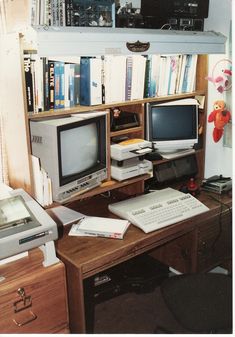 there is a desk with two computers and a printer on it in front of the bookshelf