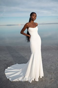 a woman standing in front of the ocean wearing a white wedding dress with long train