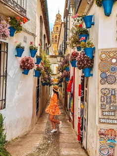 a woman in an orange dress walking down a narrow street with hanging flower pots on the buildings