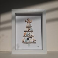 a white frame holding a small christmas tree made out of seashells and pebbles