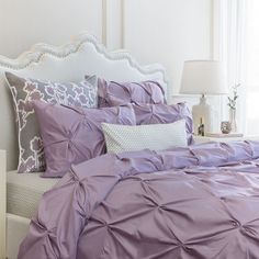 a bed with purple comforter and pillows in a white room next to a window