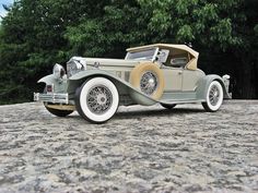 an old model car sitting on top of a stone surface next to some green trees