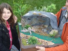 two people standing next to each other near a fake rock and grass area with plants