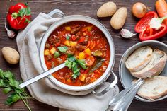 a bowl of soup with bread, tomatoes and other vegetables next to it on a wooden table