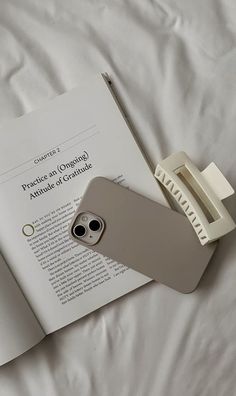 an open book and cell phone sitting on top of a white bed with the cover pulled down