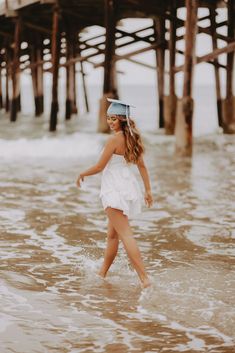 a woman in a white dress is walking through the water under a pier with her hat on