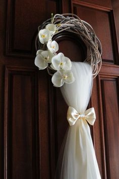 a wreath with white flowers and a bow hanging on the front door to decorate it