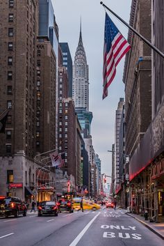 an empty city street with tall buildings in the background and american flag flying high above