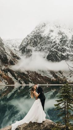 a bride and groom kissing on top of a mountain overlooking a lake with snow covered mountains in the background