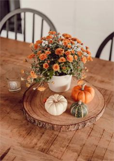 a wooden table topped with a white vase filled with orange flowers and mini pumpkins