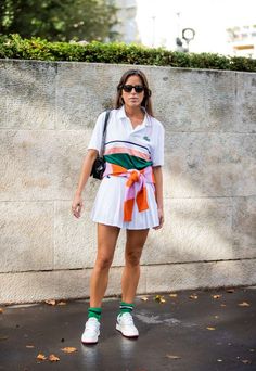 a woman standing in front of a wall wearing sunglasses and a white dress with an orange sash around her waist