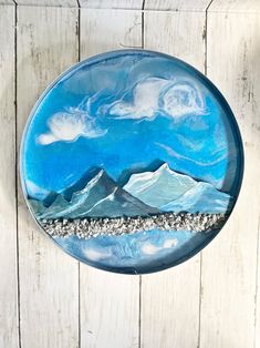 a glass plate with mountains and clouds painted on the side, sitting on a white wooden surface