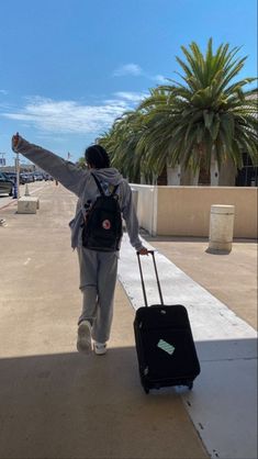 a person with a backpack and suitcase walking on the sidewalk near palm trees holding their arms in the air