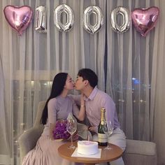 two people sitting at a table kissing each other with balloons and confetti on the wall behind them