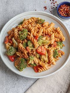 a white plate topped with pasta and broccoli next to a bowl of seasoning