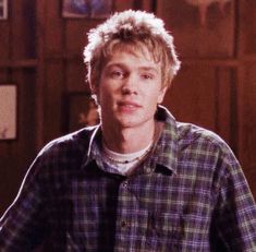 a young man standing in front of a wooden wall with pictures on the walls behind him