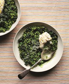 two white bowls filled with green vegetables on top of a striped table cloth next to a spoon