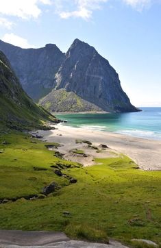 an empty beach with mountains in the background and green grass on the ground near water