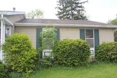 a house with green shutters in front of it and bushes on the lawn behind it