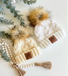 two knitted hats with pom - poms on them next to greenery