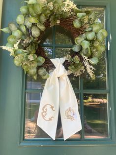 a wreath is hanging on the front door with white ribbons and green foliage, along with a monogrammed bow