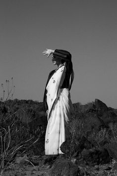 a woman standing on top of a dirt field next to rocks and plants in the desert