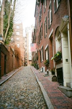 a cobblestone street lined with brick buildings and trees on both sides, in front of an american flag