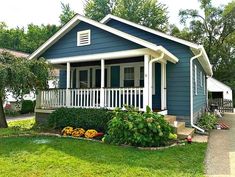 a small blue house with white porches and flowers in the front yard on a sunny day