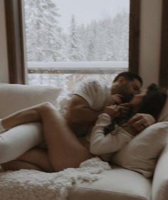 a man and woman cuddle on a couch in front of a window with snow covered trees outside