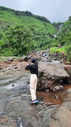 a man standing on top of a rock in the middle of a river next to a lush green hillside