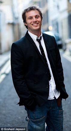 a man in a suit and tie standing on the side of an alleyway smiling at the camera