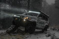 a jeep driving through the snow in front of some rocks and trees, with its lights on