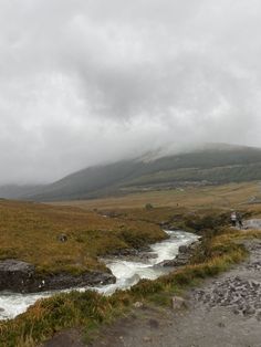 two people are standing on the edge of a stream in front of a grassy hill