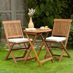 two wooden chairs sitting at a small table with white cushions on top of it in the grass