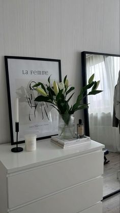 a white dresser topped with a vase filled with flowers next to a mirror and candle