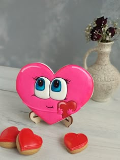 a pink heart shaped cookie with eyes on it next to some cookies and a vase