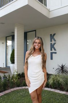 a woman standing in front of a building wearing a white dress and tan sandals with her legs crossed