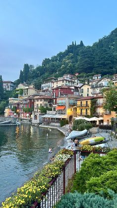 an image of a town by the water with flowers in front of it and people walking on the shore